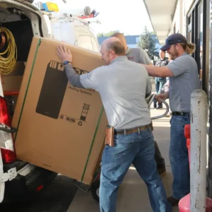 Two Elkhorn Heating Air Conditioning Plumbing and Electrical technicians loading a new Trane residential heat pump indoor unit into a branded service van for a heat pump installation in Denver Colorado