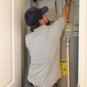 Elkhorn Heating Air Conditioning Plumbing and Electrical technician servicing the gas line and exhaust connections at the top of a residential tank water heater at a Denver Colorado home, tool belt and cordless drill visible